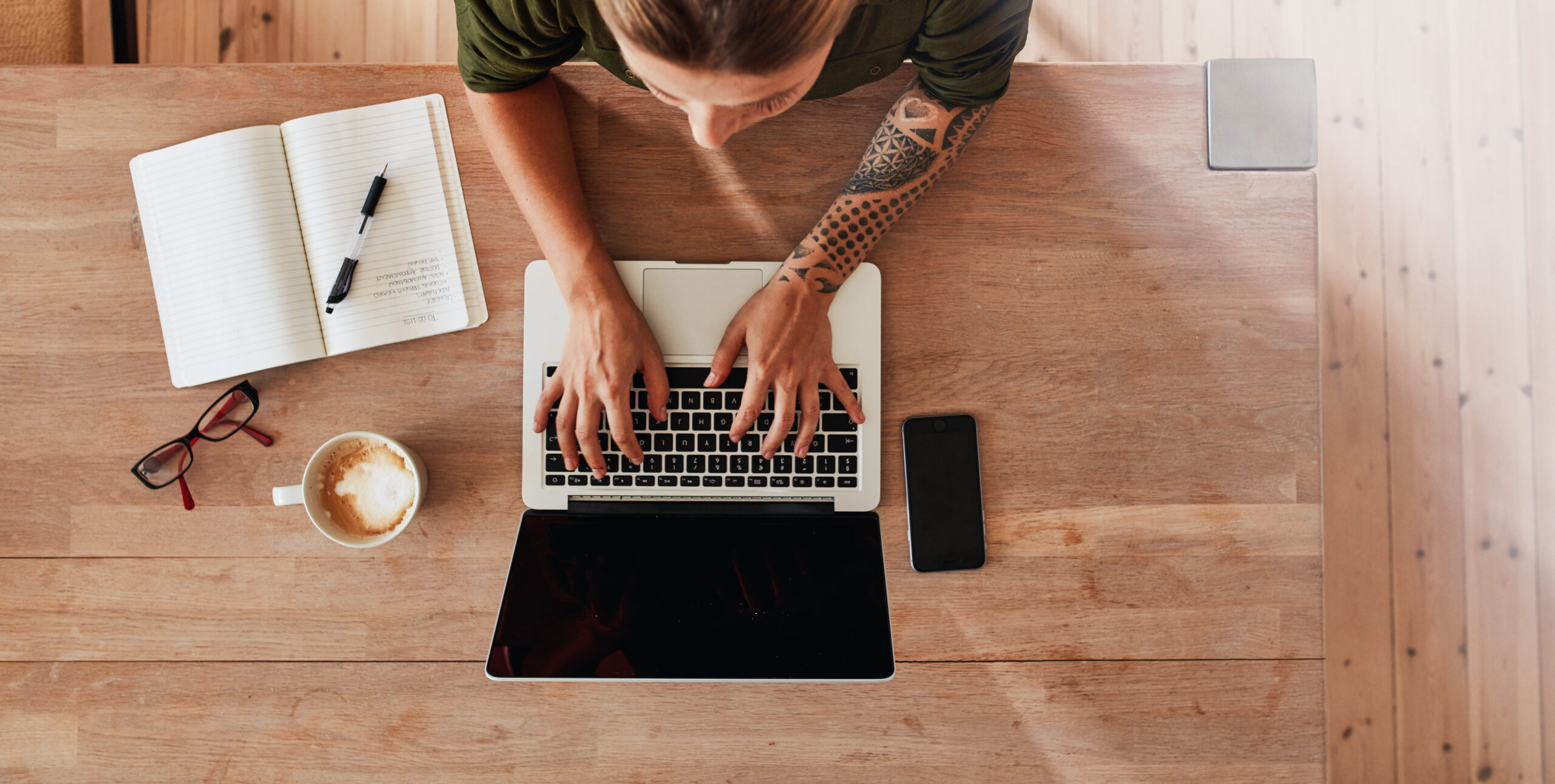 White woman with blonde hair sitting on a wooden desk typing on a computer with coffee cup, notebook, phone, pen, and reading glasses on the table.